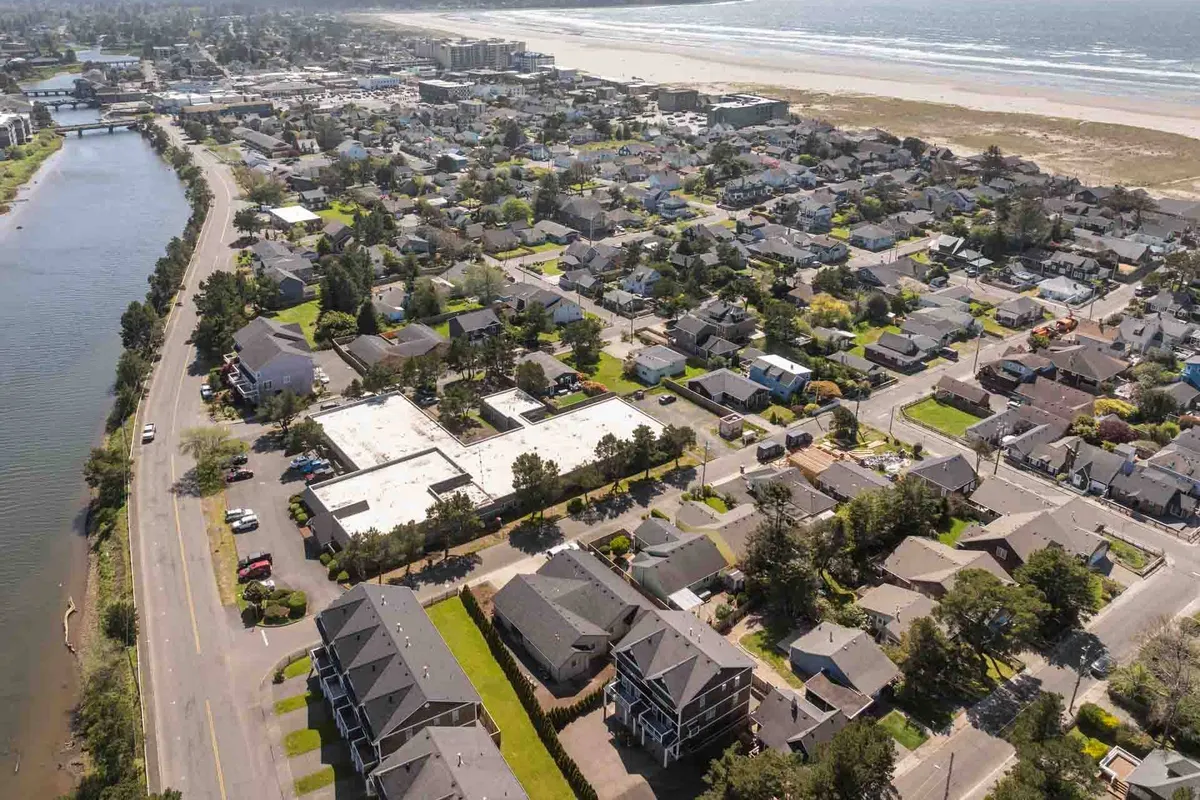 A sweeping aerial view of the coastal beach, flanked by residential homes and a river, with a commercial area visible nearby. The sandy shoreline is inviting, while the juxtaposition of houses and natural water features highlights the proximity of family-friendly activities and local attractions.