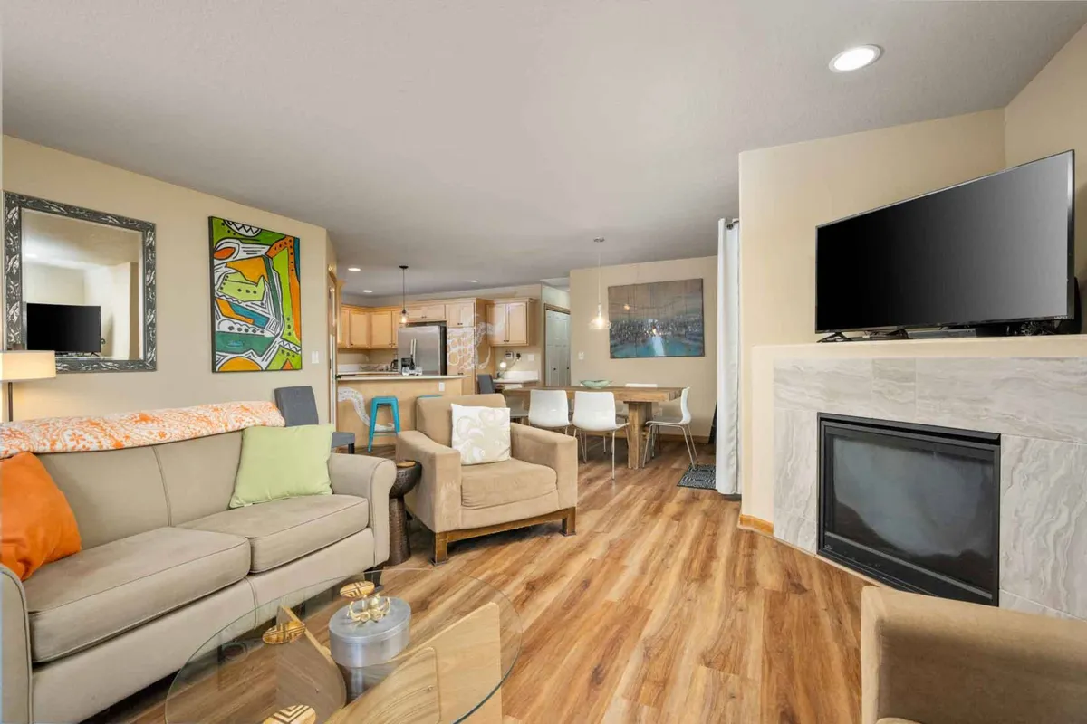 This image features a spacious open-concept living and dining area with a comfortable seating arrangement, a gas fireplace, colorful local artwork, and a dining space in the background. Natural light streams in through large windows, illuminating the modern yet inviting décor.