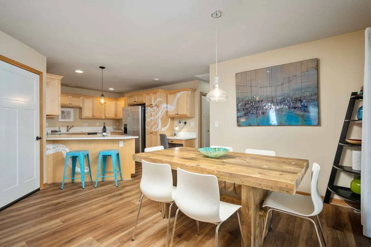 A modern dining area with a wooden table, contemporary chairs, striking local artwork on the wall, and a view toward the kitchen. Bar stools and cabinetry contribute to the open, social feel.