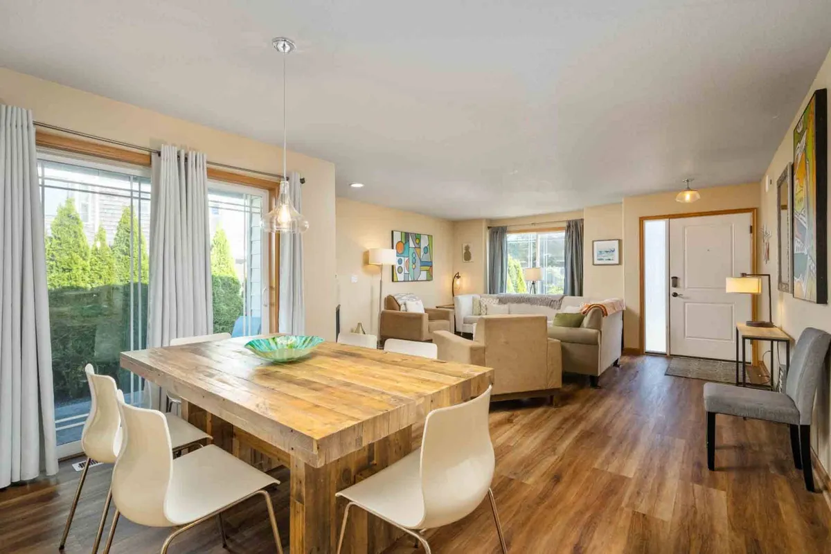 The image showcases a spacious, sunlit dining area at The Octopus' Garden featuring a beautiful wooden dining table with a green decorative bowl. Comfortable seating invites relaxation, while large windows fill the room with natural light and offer glimpses of the tranquil coastal setting.