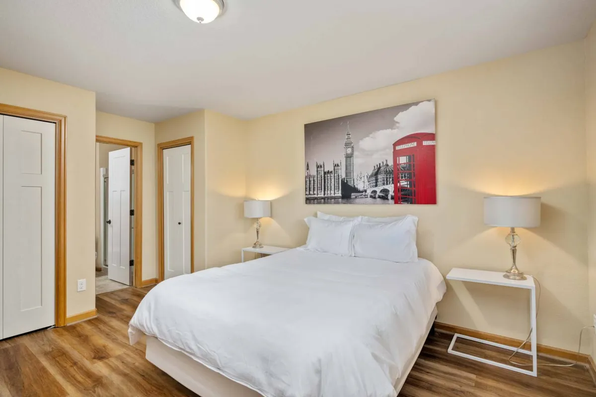 A tranquil bedroom featuring a bed with crisp white bedding, London-themed artwork above the headboard, and nightstands with soft-glowing lamps on either side. Natural light streams in from a nearby window, creating a calm and restful atmosphere ideal for relaxation and sleep.